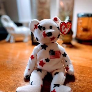 May include: A white plush bear with red and black star patterns and an American flag patch. The bear has a black nose and eyes, and a red heart-shaped tag.