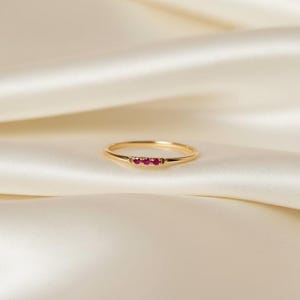 May include: A delicate gold ring with three small, vibrant red gemstones set in a row. The ring has a thin band and a polished finish, set against a soft, cream-coloured background. A minimalist piece of jewellery.