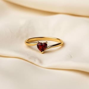 May include: A gold ring with a heart-shaped, deep red gemstone. The ring's band curves gracefully, leading to the heart-shaped stone. The ring is set against a soft, off-white background, highlighting the jewelry's design and color.