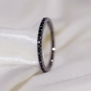 May include: A slender, black ring featuring a continuous row of small, black gemstones. The ring is displayed against a soft, white fabric, accentuating its design. This is a piece of jewellery.