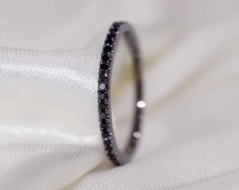 May include: A thin, black ring with a band of small, black gemstones. The ring is set against a soft, white fabric background, creating a contrast that highlights the ring's design. The ring is a piece of jewelry.