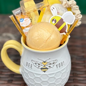 May include: A white ceramic mug with a yellow handle and rim, filled with a bath bomb, hair clip, and other yellow-themed items. The mug has a honeycomb pattern and the words "BEE YOU!" with a bee graphic.