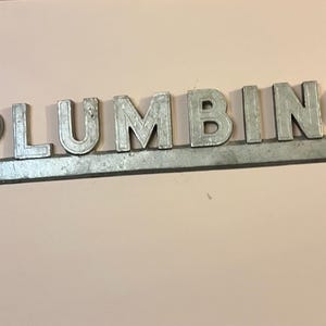 May include: A silver-colored metal sign featuring the word PLUMBING in raised, block-style capital letters. The letters are attached to a horizontal metal bar base, creating a single, cohesive piece of signage with a metallic, industrial finish.