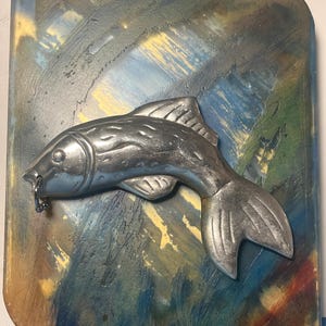 May include: A silver-colored fish sculpture with a ring in its mouth is mounted on a rectangular, multi-colored background. The background features brushstrokes of blue, green, yellow, and brown. The fish is detailed with fins and scales.
