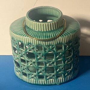 May include: A teal ceramic lantern with a woven design and a gold-colored handle. The lantern has a cylindrical shape with a ribbed top and bottom. The openwork design allows light to shine through, creating a decorative effect.