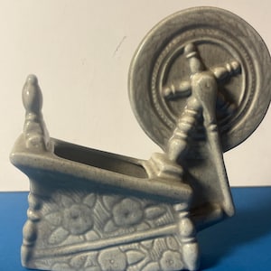 May include: A grey ceramic spinning wheel planter. The planter features a detailed design of a spinning wheel with a large wheel and a decorative base with floral patterns. The planter is a light grey colour.