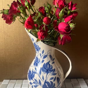 May include: A cream-colored ceramic pitcher featuring a hand-painted blue floral pattern with swirling vines and leaves. The pitcher has a curved handle and a flared spout, and it is filled with a large bouquet of vibrant, deep red roses.