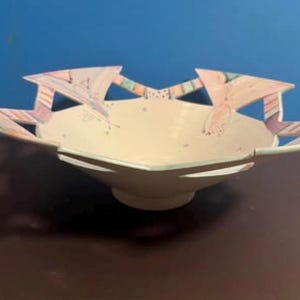 May include: A ceramic bowl with a cream-colored interior and a wide, scalloped rim featuring geometric patterns in pastel pink, blue, and purple. The bowl has a circular base and a smooth, matte finish, with distinct cut-out sections along the edge.