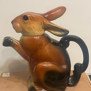 May include: A ceramic teapot shaped like a rabbit in shades of brown and tan. The rabbit has long ears, one raised paw, and a dark brown handle extending from its back. The head of the rabbit serves as a removable lid for the teapot body.