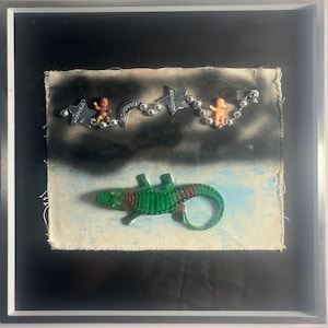 Puede incluir: Obra de arte enmarcada con un juguete de cocodrilo verde y marrón sobre un fondo texturizado. Arriba, una cadena de estrellas y cuentas plateadas conecta pequeñas figuras de bebés de plástico. El fondo tiene un efecto de cielo pintado con tonos negros y azules.