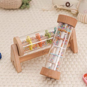 May include: Wooden baby toy with a clear tube filled with colorful beads. The toy has a wooden frame and end caps. The word "Beryl" is engraved on the side. Other wooden toys are in the background.