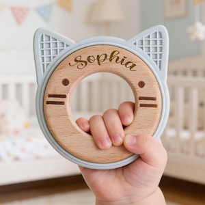 May include: A baby's teething toy in the shape of a cat's face. The toy is light blue and wood-toned, with the name "Sophia" engraved on the wooden center. The toy is held in a baby's hand. The background includes a crib and nursery decor.