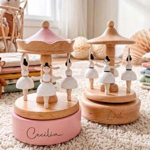 May include: Two wooden carousel music boxes featuring ballerina figures with white tutus. The left carousel has a pink base with the name Cecilia, and the right carousel has a natural wood base with the name Linda engraved on the side.