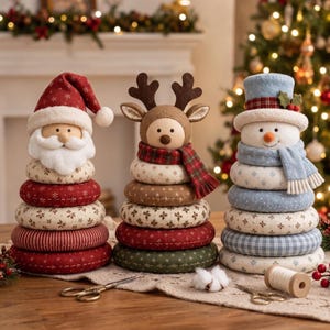 Könnte beinhalten: Drei Stoff-Stapelspielzeuge in Form von Weihnachtsmann, Rentier und Schneemann. Jede Figur besteht aus gestapelten Ringen mit verschiedenen Mustern wie Schneeflocken, Streifen und Karos in den Farben Rot, Grün, Weiß und Blau.