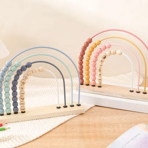 May include: Two wooden rainbow bead mazes. One has blue, green, and gray arches with the name "Eldred" spelled out in wooden beads. The other has pink, yellow, and orange arches with the name "Jamie" spelled out in wooden beads. Both sit on a wooden base.