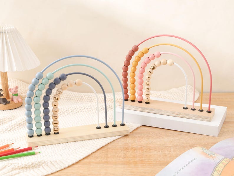 May include: Two wooden rainbow bead mazes. One has blue, green, and gray arches with the name "Eldred" spelled out in wooden beads. The other has pink, yellow, and orange arches with the name "Jamie" spelled out in wooden beads. Both sit on a wooden base.