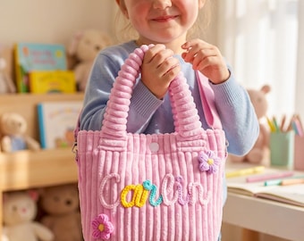 May include: A light pink corduroy tote bag featuring two rounded handles and a snap button closure. The front is decorated with the name Carlin in raised, multi-coloured yarn lettering and two small, crocheted flower appliques in pink and purple.