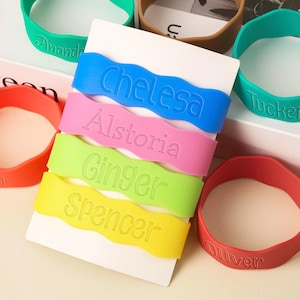 May include: A collection of colorful silicone wristbands in various colors, including blue, pink, green, and yellow. Each band features a name in a matching color. Additional bands in red, green, and brown are also visible. The bands are personalized.