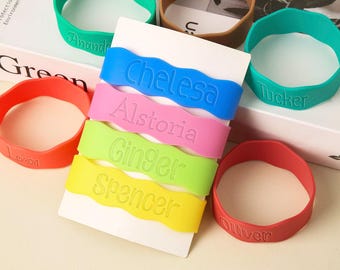 May include: A collection of colorful silicone wristbands in various colors, including blue, pink, green, and yellow. Each band features a name in a matching color. Additional bands in red, green, and brown are also visible. The bands are personalized.