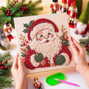 May include: A square canvas featuring a Santa Claus portrait created with small, round, colorful beads. The design includes a white beard, red hat, and green accents, surrounded by decorative sprigs of holly and berries in a beaded texture.