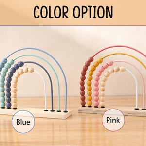 May include: Wooden rainbow abacus toys in blue and pink color options. Each abacus features arched wires with colorful beads and a wooden base. The text "COLOR OPTION" is at the top, with "Blue" and "Pink" labels below each abacus.