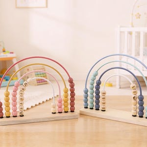 May include: Two wooden rainbow-shaped abacus toys. One has pink, yellow, and brown arches with pink and yellow beads. The other has blue, gray, and white arches with blue and gray beads. Each has a wooden base and beads with letters.