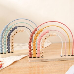 May include: Two wooden rainbow-shaped abacus toys. One features pastel blue, green, and gray arches with the name "Gordon" on the beads. The other has pink, yellow, and white arches with the name "Roisin". Each sits on a wooden base.