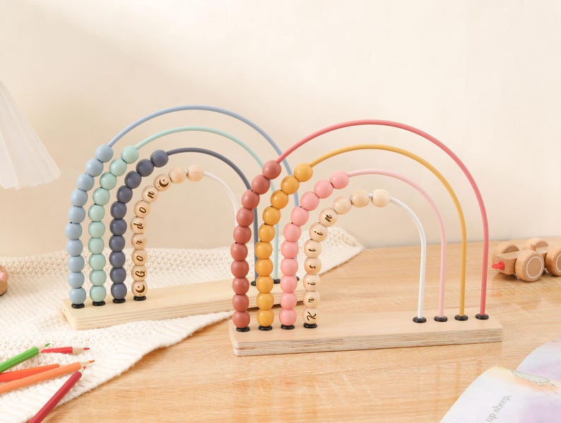 May include: Two wooden rainbow-shaped abacus toys. One features pastel blue, green, and gray arches with the name "Gordon" on the beads. The other has pink, yellow, and white arches with the name "Roisin". Each sits on a wooden base.