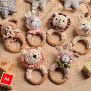 May include: A collection of handmade crochet baby rattles in the shape of animals, including a bear, lion, deer, and giraffe. Each rattle has a wooden ring and is in neutral colors. Wooden blocks with letters and images are also present.