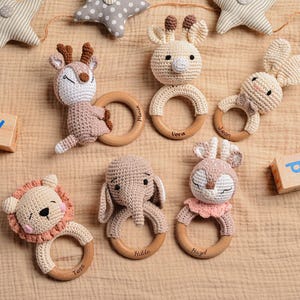 May include: A collection of handmade crochet baby rattles in the shape of animals, including a deer, giraffe, bunny, lion, elephant, and a deer. Each rattle has a wooden ring and is in neutral colors.