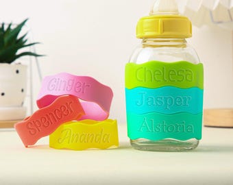 Peut inclure: Une bouteille pour bébé transparente avec un bouchon jaune et une housse en silicone verte, bleue et jaune. La housse porte les noms Chelesa, Jasper et Alstoria. Il y a aussi quatre bracelets en silicone avec les noms Ginger, Spencer et Ananda.