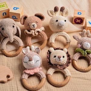 May include: A collection of handmade crochet baby rattles in the shape of animals, including an elephant, bear, giraffe, deer, lion, and bunny. Each rattle has a wooden ring and is in neutral colors. Wooden blocks with letters are in the background.