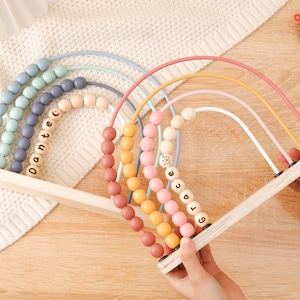 May include: A wooden rainbow abacus toy with colorful beads and the names "Dante" and "Grace" spelled out. The abacus has a light wood frame and is held by a person. The beads are in shades of blue, pink, orange, and yellow.