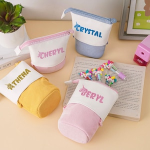 May include: A collection of personalized pencil cases in various colors, including yellow, pink, and blue. Each case features a name embroidered in a contrasting color. The cases are made of a textured fabric and are displayed with unicorn-themed accessories.