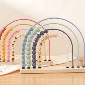May include: Wooden rainbow-shaped bead mazes in pastel colors. Each arch has a different color and wooden beads. The names "Madge" and "Fabian" are spelled out with wooden letter beads. A wooden toy car is in the background.