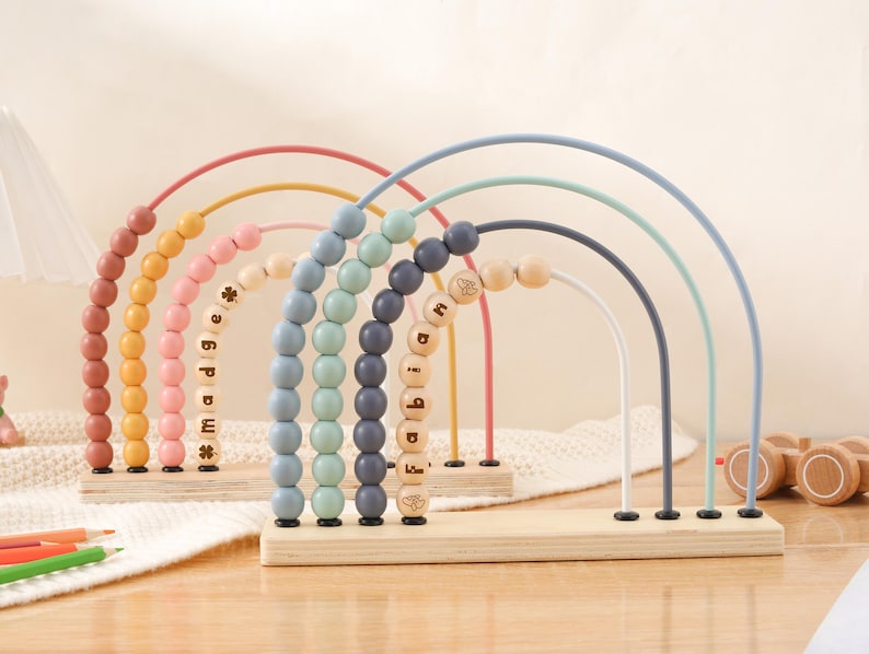 May include: Wooden rainbow-shaped bead mazes in pastel colors. Each arch has a different color and wooden beads. The names "Madge" and "Fabian" are spelled out with wooden letter beads. A wooden toy car is in the background.
