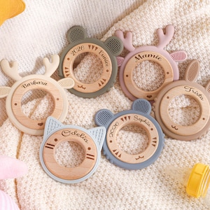 May include: A collection of baby teethers in various pastel colors, each with a wooden center and a unique animal-themed design. The teethers feature names like "Barbara" and "Sophia" and the year "2026".