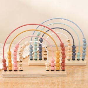 May include: Wooden rainbow abacus toy with colorful beads and arched wires. The abacus has a wooden base and the beads are in shades of pink, yellow, orange, blue, and green. The name "Norma" is spelled out on the beads of one abacus.