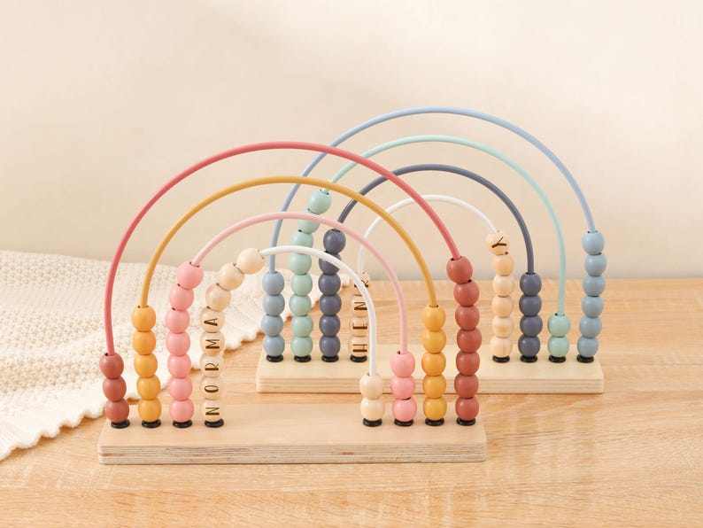 May include: Wooden rainbow abacus toy with colorful beads and arched wires. The abacus has a wooden base and the beads are in shades of pink, yellow, orange, blue, and green. The name "Norma" is spelled out on the beads of one abacus.