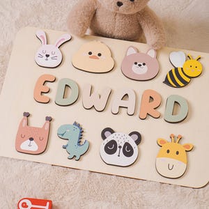 May include: A wooden name puzzle with the name "EDWARD" spelled out in colorful letters. The puzzle includes animal-shaped pieces like a bunny, duck, bear, bee, fox, dinosaur, panda, and giraffe. A teddy bear sits above the puzzle.