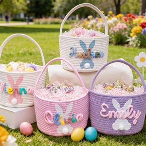 以下が含まれることがあります： 白、ピンク、紫の4つのイースターバスケット。それぞれにウサギのデザインと名前がついています。バスケットにはカラフルな卵と装飾用の草が詰められ、花が咲き乱れる芝生の上に置かれています。