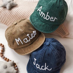 May include: Three personalized baseball caps in shades of green, brown, and blue. Each cap features a name embroidered in white: "Amber," "Mateo," and "Jack." The caps are arranged on a light-colored surface.