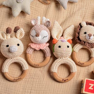 May include: A collection of handmade crochet baby rattles in the shape of animals, including a lion, bunny, and deer. Each rattle has a wooden ring and is personalized with a name. The toys are beige, brown, and pink.