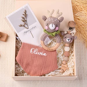 May include: A baby gift set in a box includes a crochet deer rattle, a pacifier clip, and a bib embroidered with "Olivia." A card with dried flowers and wooden blocks are also included. The box is filled with shredded paper.
