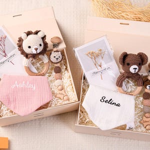 May include: Two baby gift boxes with a lion and bear theme. Each box contains a crochet rattle, pacifier clip, bib with a name, and a card. The bibs are pink and white. The rattles and clips are brown and beige.