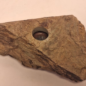 Könnte beinhalten: Ein Kerzenhalter aus Naturstein mit einer kreisförmigen Öffnung. Der Stein ist eine Mischung aus Braun- und Beigetönen mit sichtbarer Textur und Maserung. Der Kerzenhalter ist für eine kleine Kerze konzipiert.