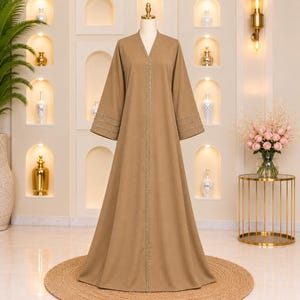 May include: A long-sleeved, floor-length tan abaya featuring a V-neckline and a central vertical trim of silver embellishments. The wide sleeves are accented with three horizontal rows of matching silver detailing at the cuffs.