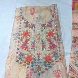 Puede incluir: Túnica bordada de color crema con motivos florales en rosa, azul y naranja. El escote presenta intrincados bordados con lentejuelas. La tela tiene una calidad transparente y el diseño incluye delicados motivos florales.