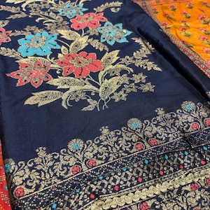 以下が含まれることがあります： 紺色の生地に金色の花の刺繍。赤、青、金色の大きな花がデザインされています。赤と青のアクセントが付いた詳細な金のボーダーが下部にあります。この生地は、衣類や室内装飾に使用される可能性があります。