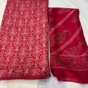 May include: A two-piece set of red fabric with white ornate patterns. One piece is folded, showing a scalloped edge. The other piece is a sheer fabric with diagonal stripes and floral designs. The set is likely for clothing or textiles.